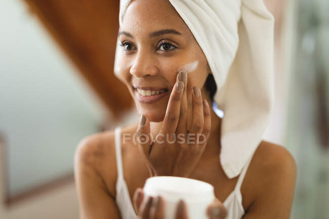 Femme de race mixte dans la salle de bain appliquant crème visage pour les soins de la peau, souriant. mode de vie domestique, profiter de loisirs d'auto-soins à la maison. — Photo de stock