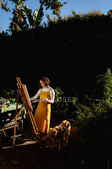 Белая женщина в солнечном саду со своей собакой, рисующая на холсте. домашний образ жизни, наслаждаясь отдыхом дома. — стоковое фото