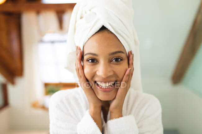 Портрет улыбающейся женщины смешанной расы в ванной комнате, держащей лицо после увлажнения, смотрящей в камеру. — стоковое фото
