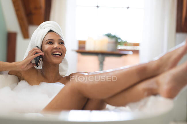 Lachende Mischlingshündin im Badezimmer, die mit erhobenen Füßen auf dem Smartphone spricht. häuslicher Lebensstil, selbstgepflegte Freizeit zu Hause genießen. — Stockfoto