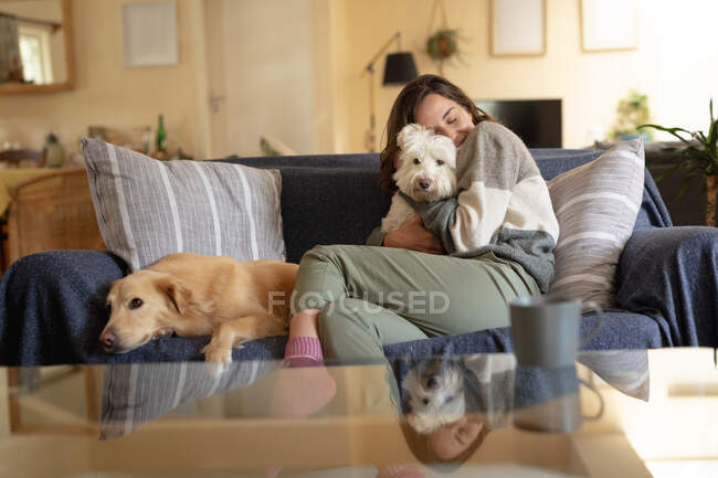 Улыбающаяся белая женщина в гостиной, сидящая на диване и обнимающая свою собаку. домашний образ жизни, наслаждаясь отдыхом дома. — стоковое фото