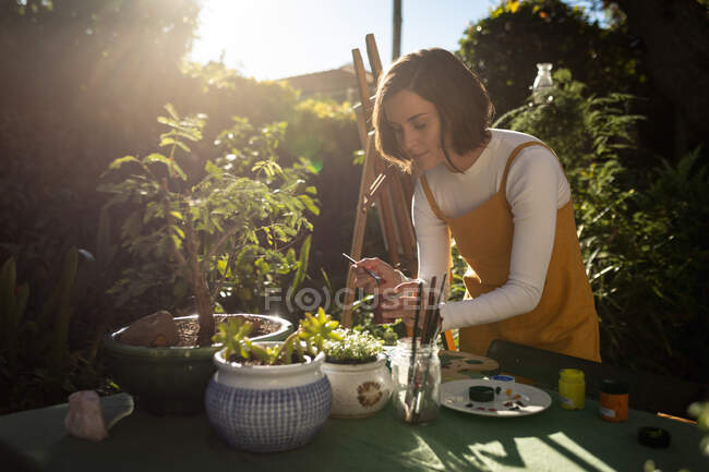 Белая женщина в солнечном саду, рисует, смешивает краску. домашний образ жизни, наслаждаясь отдыхом дома. — стоковое фото