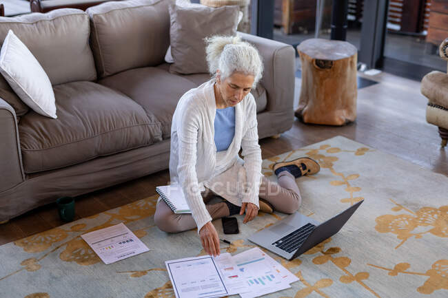 Femme caucasienne senior dans le salon assis sur le sol et travaillant sur ordinateur portable. mode de vie à la retraite, passer du temps seul à la maison. — Photo de stock