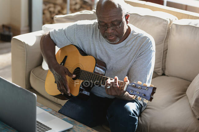 Homem americano africano sênior na moderna sala de estar situada no sofá e tocando violão. estilo de vida aposentadoria, passar o tempo sozinho em casa. — Fotografia de Stock