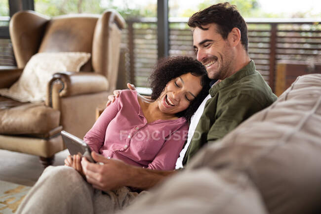 Felice coppia diversificata seduta sul divano in soggiorno utilizzando tablet. trascorrendo del tempo a casa in un appartamento moderno. — Foto stock