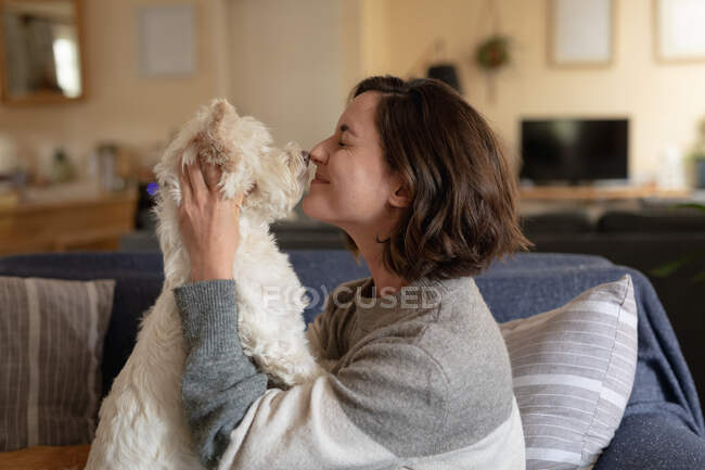 Smiling caucasian woman in living room sitting on sofa embracing her pet dog. domestic lifestyle, enjoying leisure time at home. — Stock Photo