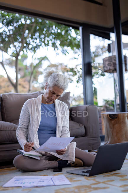 Donna caucasica anziana in soggiorno seduta sul pavimento e al lavoro. stile di vita di pensione, trascorrere del tempo da solo a casa. — Foto stock