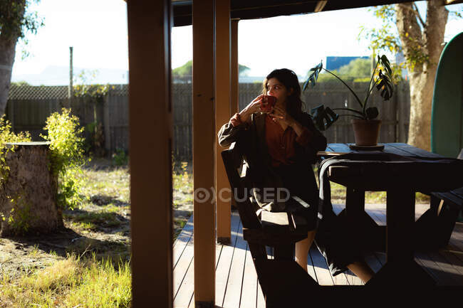 Mixed race woman sitting and drinking coffee in sunny garden. healthy lifestyle, enjoying leisure time at home. — Stock Photo