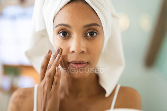 Mujer de raza mixta en baño aplicando crema facial para el cuidado de la piel. estilo de vida doméstico, disfrutando del tiempo libre de autocuidado en casa. - foto de stock