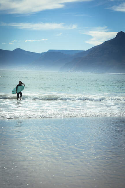 Mujer de raza mixta caminando y sosteniendo tabla de surf en el mar en el día soleado. estilo de vida saludable, disfrutar del tiempo libre al aire libre. - foto de stock