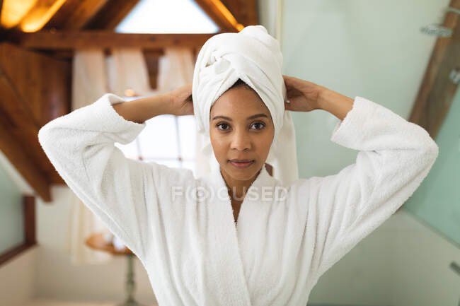 Porträt einer lächelnden Mischlingsfrau im Badezimmer, die sich ein Handtuch auf den Kopf bindet und in die Kamera blickt. häuslicher Lebensstil, selbstgepflegte Freizeit zu Hause genießen. — Stockfoto