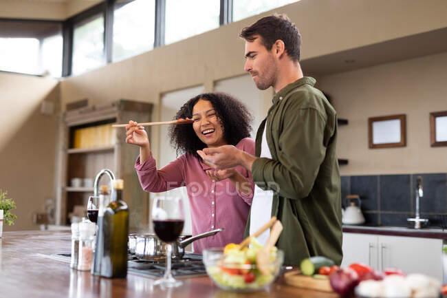 Feliz casal diversificado na cozinha preparando comida juntos comendo e sorrindo. passar o tempo fora em casa no apartamento moderno. — Fotografia de Stock