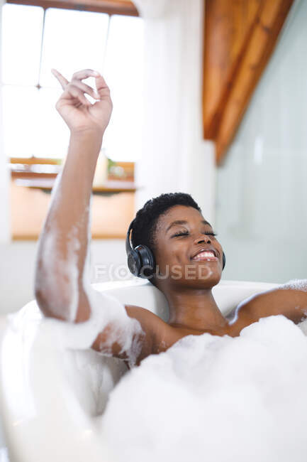 Усміхнена афро-американська жінка відпочиває у ванній з закритими очима, слухаючи музику на навушниках.. — стокове фото