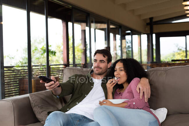 Feliz casal diversificado sentado no sofá na sala de estar abraçando e assistindo tv. passar o tempo fora em casa no apartamento moderno. — Fotografia de Stock