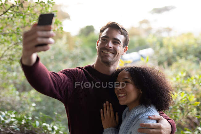 Felice coppia diversificata sul balcone abbracciando prendendo selfie e sorridente. trascorrere del tempo libero a casa. — Foto stock
