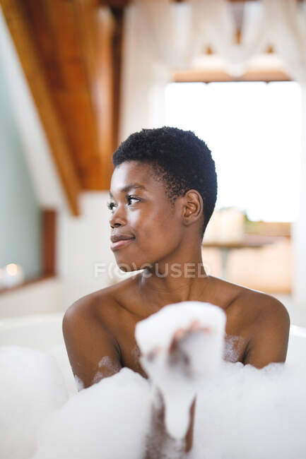 Femme afro-américaine souriante dans la salle de bain relaxante dans un bain en mousse. mode de vie domestique, profiter de loisirs d'auto-soins à la maison. — Photo de stock