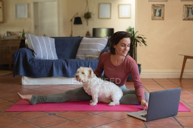 Femme caucasienne souriante dans le salon avec son chien de compagnie, pratiquer le yoga, en utilisant un ordinateur portable. mode de vie domestique, profiter du temps libre à la maison. — Photo de stock