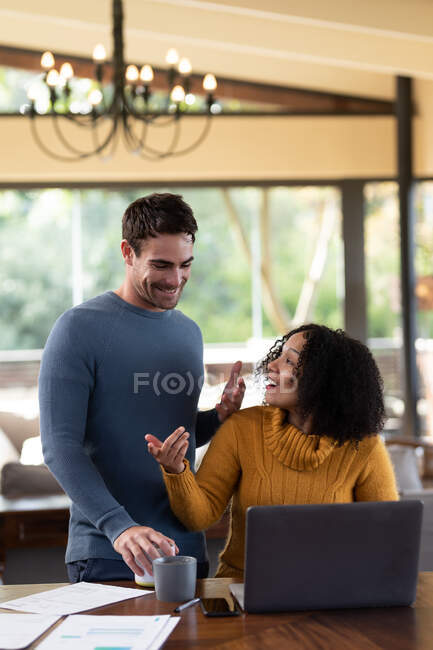 Casal diverso feliz sentado à mesa sorrindo e usando laptop. trabalhando em casa no apartamento moderno. — Fotografia de Stock