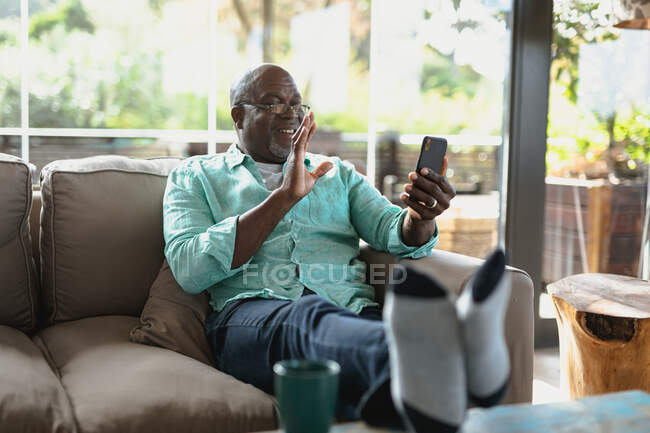 Heureux homme afro-américain âgé assis sur le canapé et faire un appel vidéo dans le salon moderne. mode de vie à la retraite, passer du temps seul à la maison. — Photo de stock