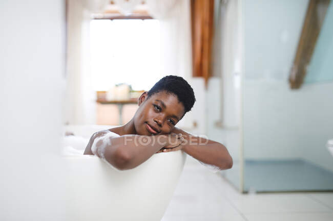 Porträt einer lächelnden afrikanisch-amerikanischen Frau im Badezimmer, die sich in der Badewanne entspannt. häuslicher Lebensstil, selbstgepflegte Freizeit zu Hause genießen. — Stockfoto