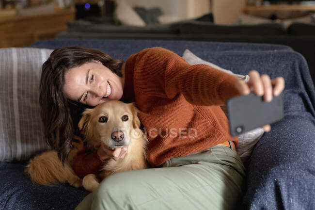 Усміхнена біла жінка у вітальні, сидячи на дивані, обіймає свого домашнього собаку, приймаючи селфі. домашній спосіб життя, насолоджуючись дозвіллям вдома . — стокове фото