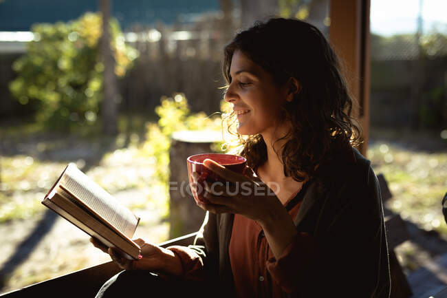 Mujer de raza mixta leyendo libro y tomando café en un jardín soleado. estilo de vida saludable, disfrutando del tiempo libre en casa. - foto de stock