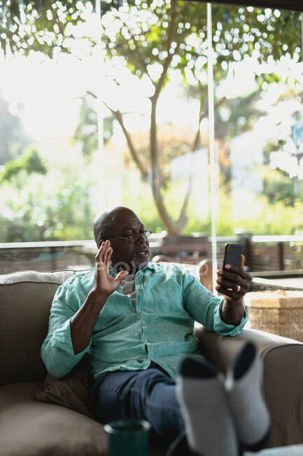 Homme afro-américain senior assis sur le canapé et faire un appel vidéo dans le salon moderne. mode de vie à la retraite, passer du temps seul à la maison. — Photo de stock