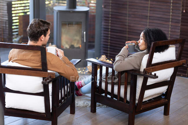 Feliz casal diversificado na sala de estar sentado à lareira segurando canecas e beber café. passar o tempo fora em casa. . — Fotografia de Stock
