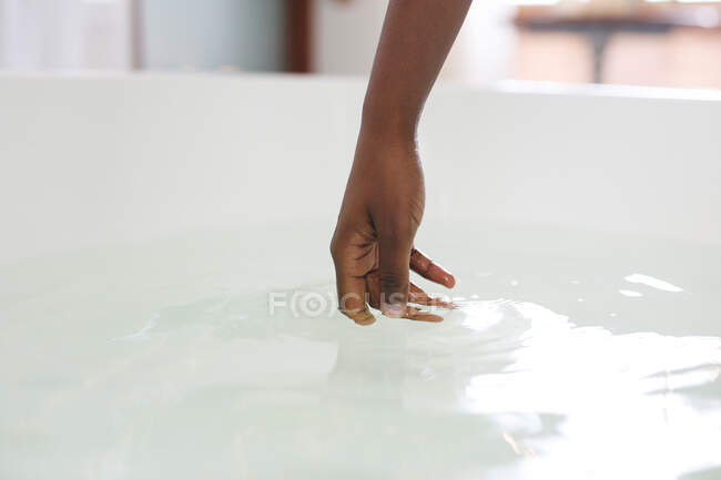 Gros plan de la main de la femme afro-américaine dans la salle de bain touchant l'eau dans le bain. mode de vie domestique, profiter de loisirs d'auto-soins à la maison. — Photo de stock