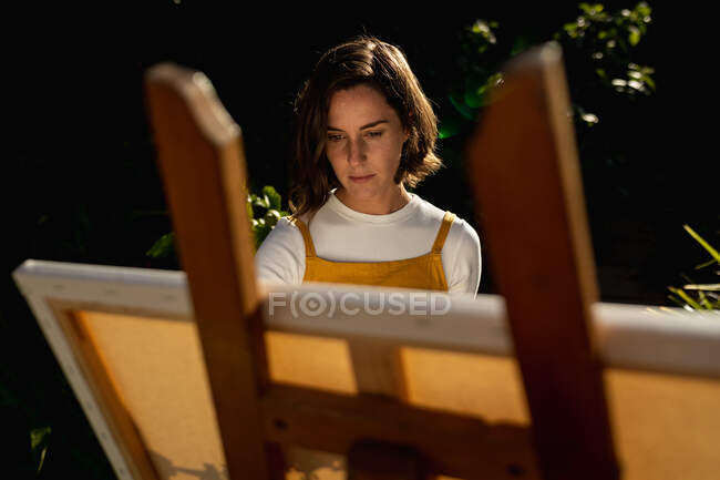 Mujer caucásica en jardín soleado, pintando sobre lienzo. estilo de vida doméstico, disfrutando del tiempo libre en casa. - foto de stock