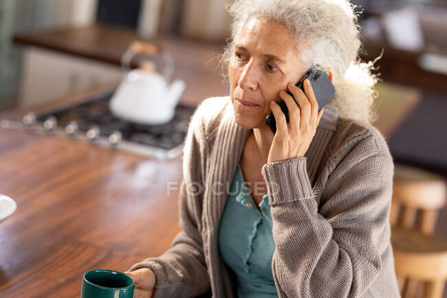 Eine ältere kaukasische Frau entspannt in der Küche mit dem Smartphone und trinkt Kaffee. Lebensstil im Ruhestand, Zeit allein zu Hause verbringen. — Stockfoto