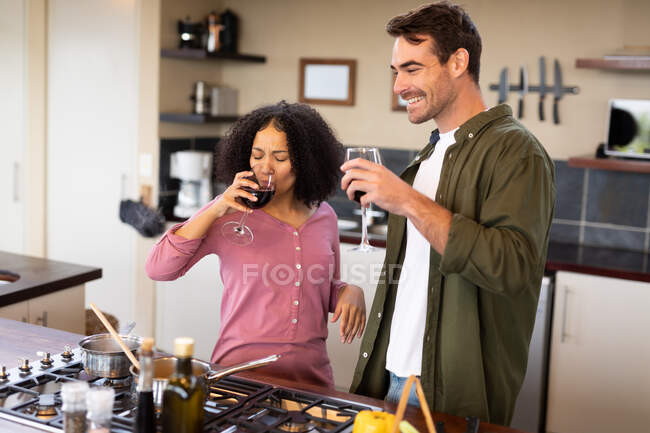 Feliz pareja diversa en la cocina preparando la comida juntos bebiendo vino. pasar tiempo libre en casa en apartamento moderno. - foto de stock