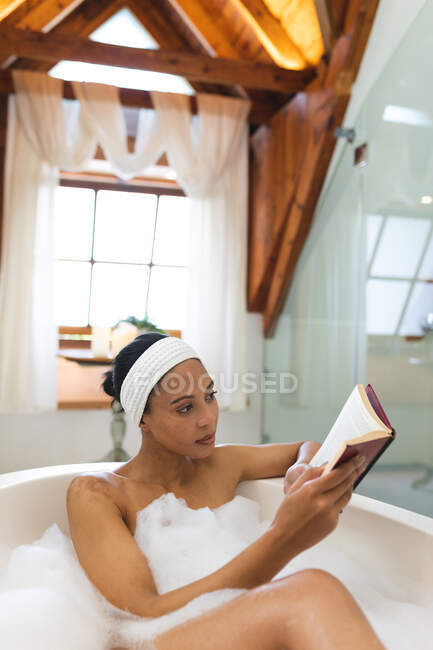 Змішана расова жінка у ванній кімнаті, розслабляється у ванній читаючи книгу. домашній спосіб життя, насолоджуючись доглядом за дозвіллям вдома . — стокове фото