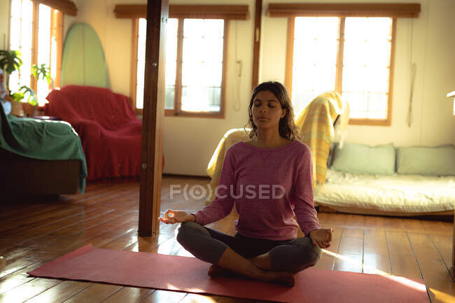 Femme mixte pratiquant le yoga, assise en train de méditer dans le salon ensoleillé. mode de vie sain, profiter de loisirs à la maison. — Photo de stock