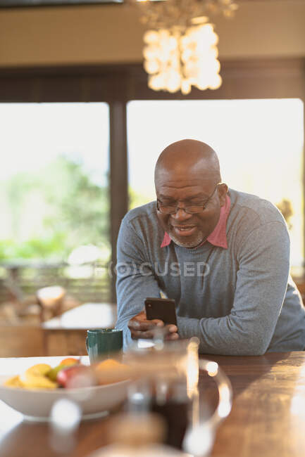 Glücklicher älterer afrikanisch-amerikanischer Mann, der in der modernen Küche steht und das Smartphone benutzt. Lebensstil im Ruhestand, Zeit allein zu Hause verbringen. — Stockfoto