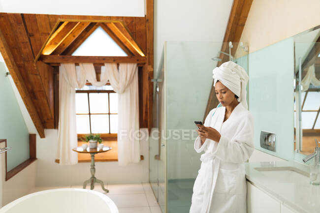 Змішана расова жінка у ванній кімнаті за допомогою смартфона та чищення зубів. домашній спосіб життя, насолоджуючись доглядом за дозвіллям вдома . — стокове фото