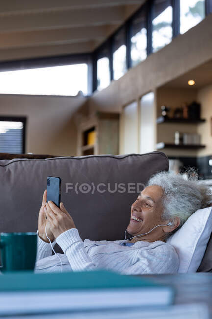 Glückliche Seniorin, die im modernen Wohnzimmer liegt und das Smartphone benutzt. Lebensstil im Ruhestand, Zeit allein zu Hause verbringen. — Stockfoto