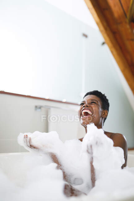 Feliz mujer afroamericana riendo en el baño, divirtiéndose con espuma de baño. estilo de vida doméstico, disfrutando del tiempo libre de autocuidado en casa. - foto de stock
