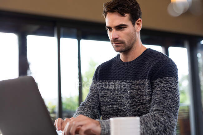 Homem caucasiano sentado à mesa na cozinha trabalhando remotamente usando laptop. passar o tempo fora em casa no apartamento moderno. — Fotografia de Stock
