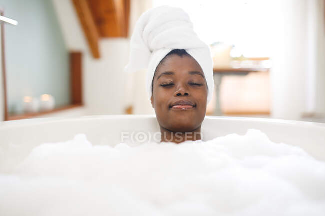 Femme afro-américaine souriante dans la salle de bain relaxant dans la baignoire avec les yeux fermés. mode de vie domestique, profiter de loisirs d'auto-soins à la maison. — Photo de stock