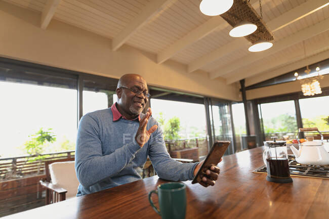 Lächelnder älterer afrikanisch-amerikanischer Mann sitzt in der modernen Küche und telefoniert per Video. Lebensstil im Ruhestand, Zeit allein zu Hause verbringen. — Stockfoto