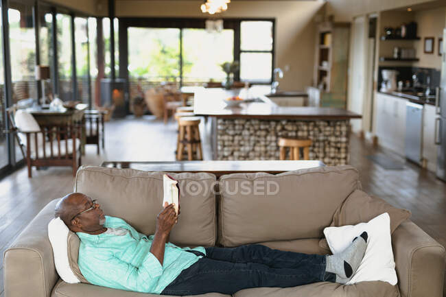 Relaxante homem afro-americano sênior deitado no sofá e lendo livro na sala de estar moderna. estilo de vida aposentadoria, passar o tempo sozinho em casa. — Fotografia de Stock