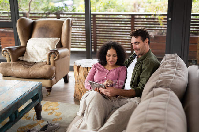 Feliz pareja diversa sentado en el sofá en la sala de estar utilizando la tableta. pasar tiempo libre en casa en apartamento moderno. - foto de stock