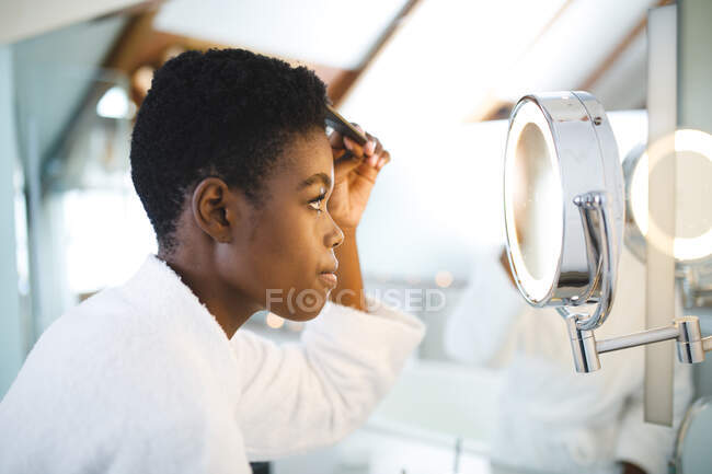 Mujer afroamericana en el baño, mirándose en el espejo y peinándose el pelo. estilo de vida doméstico, disfrutando del tiempo libre de autocuidado en casa. - foto de stock