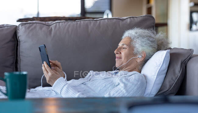 Mujer caucásica mayor que pone y utiliza el teléfono inteligente en la sala de estar moderna. estilo de vida de jubilación, pasar tiempo solo en casa. - foto de stock