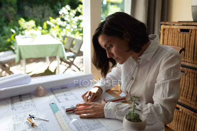 Architecte caucasienne dans le salon, assise à la table de travail, plans de dessin. mode de vie domestique, travail à distance de la maison. — Photo de stock