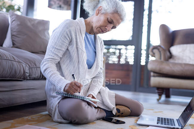Femme caucasienne âgée dans le salon assis sur le sol et écrivant. mode de vie à la retraite, passer du temps seul à la maison. — Photo de stock
