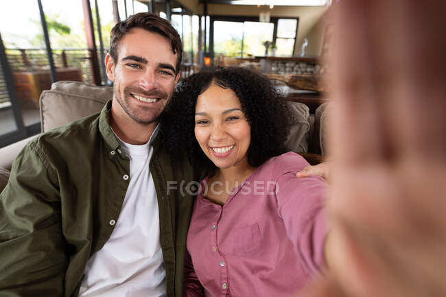 Retrato de feliz pareja diversa sentada en el sofá en la sala de estar tomando selfie y sonriendo. pasar tiempo libre en casa en apartamento moderno. - foto de stock