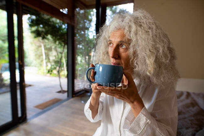 Thoughtful senior caucasian woman sitting on the bad and drinking coffee. retirement lifestyle, spending time alone at home. — Stock Photo