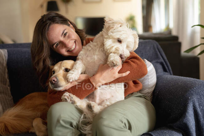 Retrato de una mujer caucásica sonriente en la sala de estar sentada en el sofá abrazando a sus perros de compañía. estilo de vida doméstico, disfrutando del tiempo libre en casa. - foto de stock